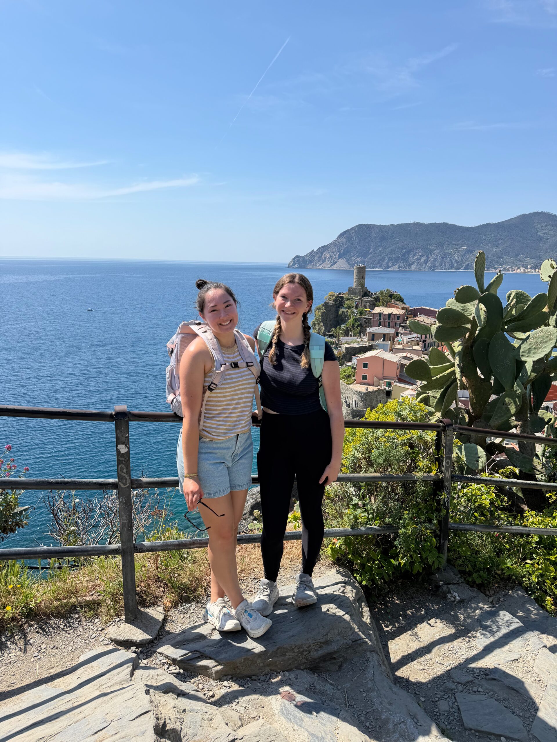 Two women stand together on a sunny coastal overlook in Cinque Terre, Italy, smiling at the camera with the blue Mediterranean Sea, colorful hillside buildings, and green mountains in the background. A large cactus and stone pathway frame the scenic view.